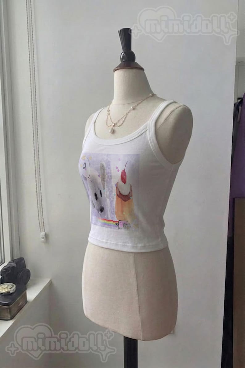 May include: A white sleeveless crop top featuring a graphic print of a white creature, a cupcake with a cherry on top, and a small cat on a rainbow. The top has a scoop neckline and is displayed on a mannequin wearing a delicate pearl necklace.