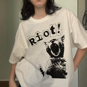 Pode incluir: Camiseta branca com um desenho gr&aacute;fico preto de um gato gritando e a palavra "Riot!" impressa acima. O design &eacute; arrojado e chamativo, ideal para uso casual.