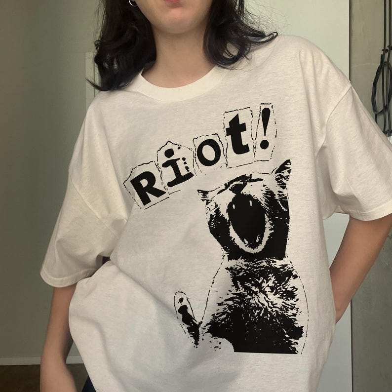Pode incluir: Camiseta branca com um desenho gr&aacute;fico preto de um gato gritando e a palavra "Riot!" impressa acima. O design &eacute; arrojado e chamativo, ideal para uso casual.