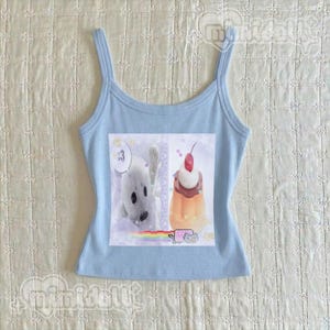 May include: A light blue ribbed tank top featuring a central graphic print. The print displays a white seal with a speech bubble containing the text :3, a pudding cup topped with a cherry, a small gray cat, and a rainbow streak across the bottom.