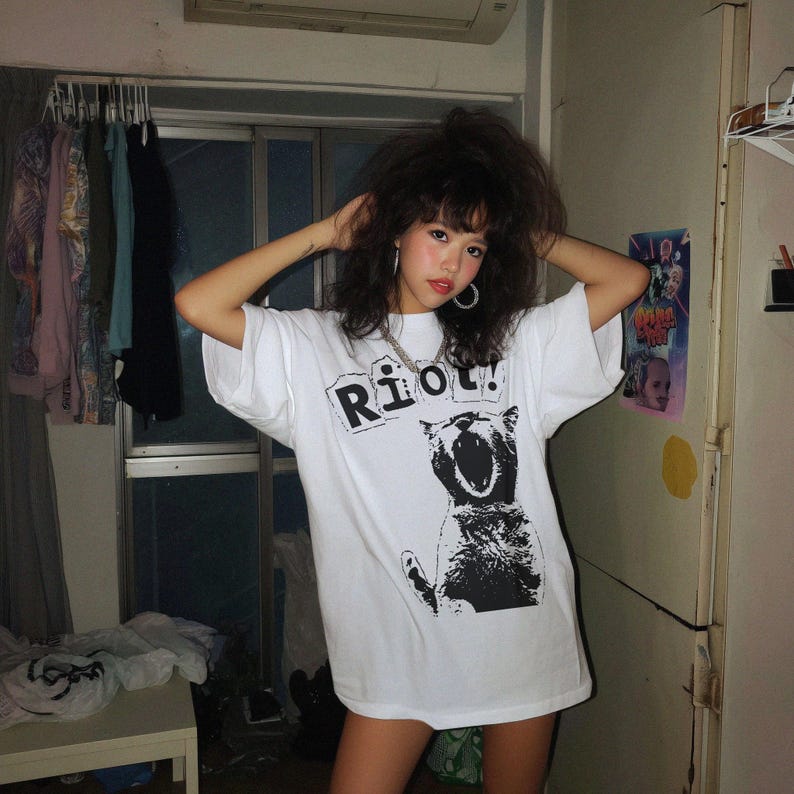 Pode incluir: T-shirt branco com a palavra "Riot!" em preto, sobre um gr&aacute;fico a preto e branco de um gato a rugir. A t-shirt tem mangas curtas e um corte folgado. Design gr&aacute;fico casual.