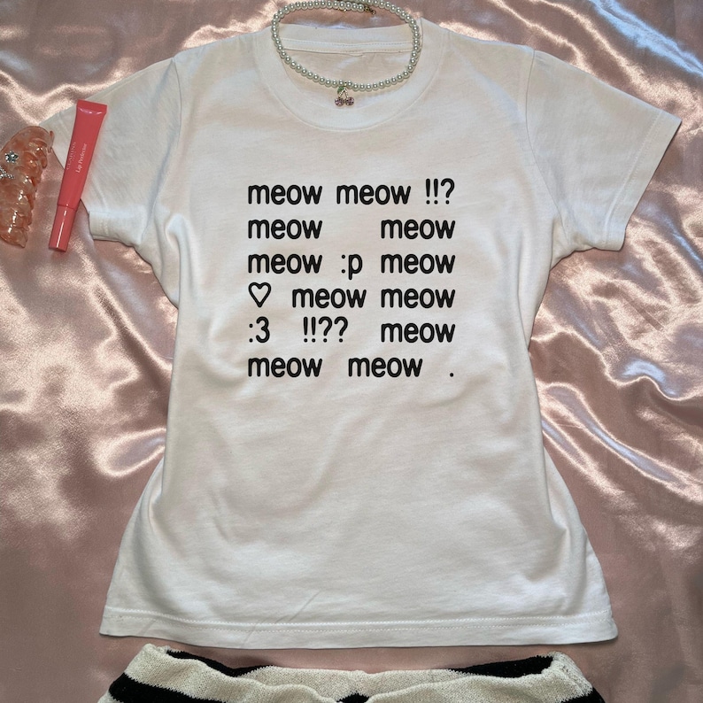 May include: White t-shirt with the text "meow meow !!? meow meow meow :p meow ♡ meow meow :3 !!?? meow meow meow ." A pearl necklace with a cherry pendant and a tube of lip gloss are also visible.