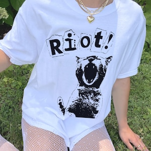 May include: White t-shirt with a black graphic of a screaming cat and the word "Riot!" in a distressed font. The shirt is made of a soft material and is a casual style. The shirt is a unisex design.
