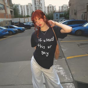 May include: Black t-shirt with the text "if u read this ur gay" in white. The person is wearing light-colored pants and a brown shoulder bag. The background shows a parking lot with cars and buildings.