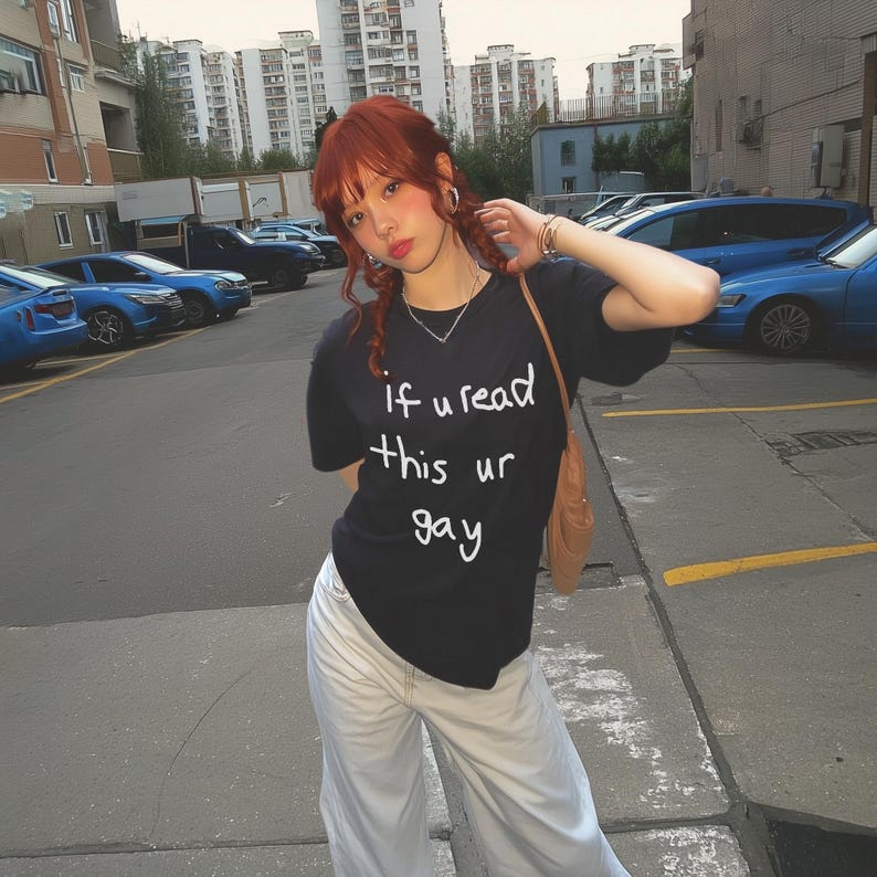 May include: Black t-shirt with the text "if u read this ur gay" in white. The person is wearing light-colored pants and a brown shoulder bag. The background shows a parking lot with cars and buildings.