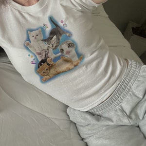 Peut inclure: T-shirt blanche courte avec un imprim&eacute; graphique de cinq chatons sur un fond bleu. Les chatons sont dans diff&eacute;rentes poses, et il y a de petits c&oelig;urs, des &eacute;toiles et des empreintes de pattes autour d'eux.