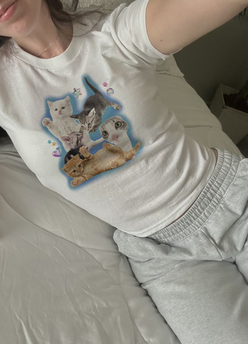 Peut inclure: T-shirt blanche courte avec un imprim&eacute; graphique de cinq chatons sur un fond bleu. Les chatons sont dans diff&eacute;rentes poses, et il y a de petits c&oelig;urs, des &eacute;toiles et des empreintes de pattes autour d'eux.