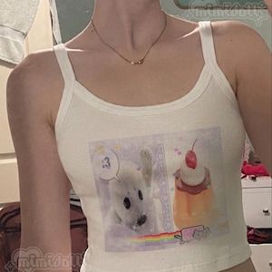 May include: A cream-colored ribbed tank top featuring a graphic print on the front. The print displays a white seal pup, a custard dessert topped with a cherry, and a small pixelated cat with a rainbow trail. The text 13 appears in a speech bubble.