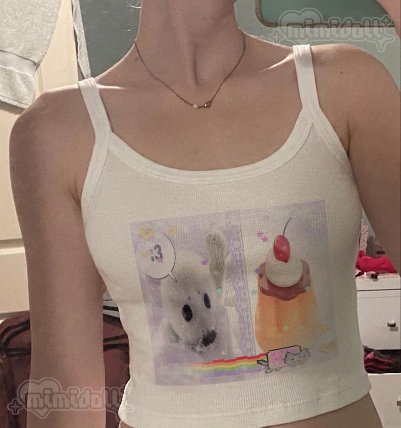 May include: A cream-colored ribbed tank top featuring a graphic print on the front. The print displays a white seal pup, a custard dessert topped with a cherry, and a small pixelated cat with a rainbow trail. The text 13 appears in a speech bubble.