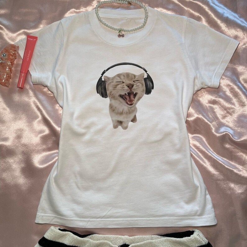 May include: White t-shirt with a graphic of a cat wearing headphones and singing. The cat is light brown and white with black headphones.