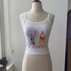 May include: A white sleeveless tank top featuring a graphic print of a white seal with a speech bubble containing the number 3, a dessert topped with a cherry, and a small rainbow. The tank top is displayed on a mannequin wearing a pearl necklace.