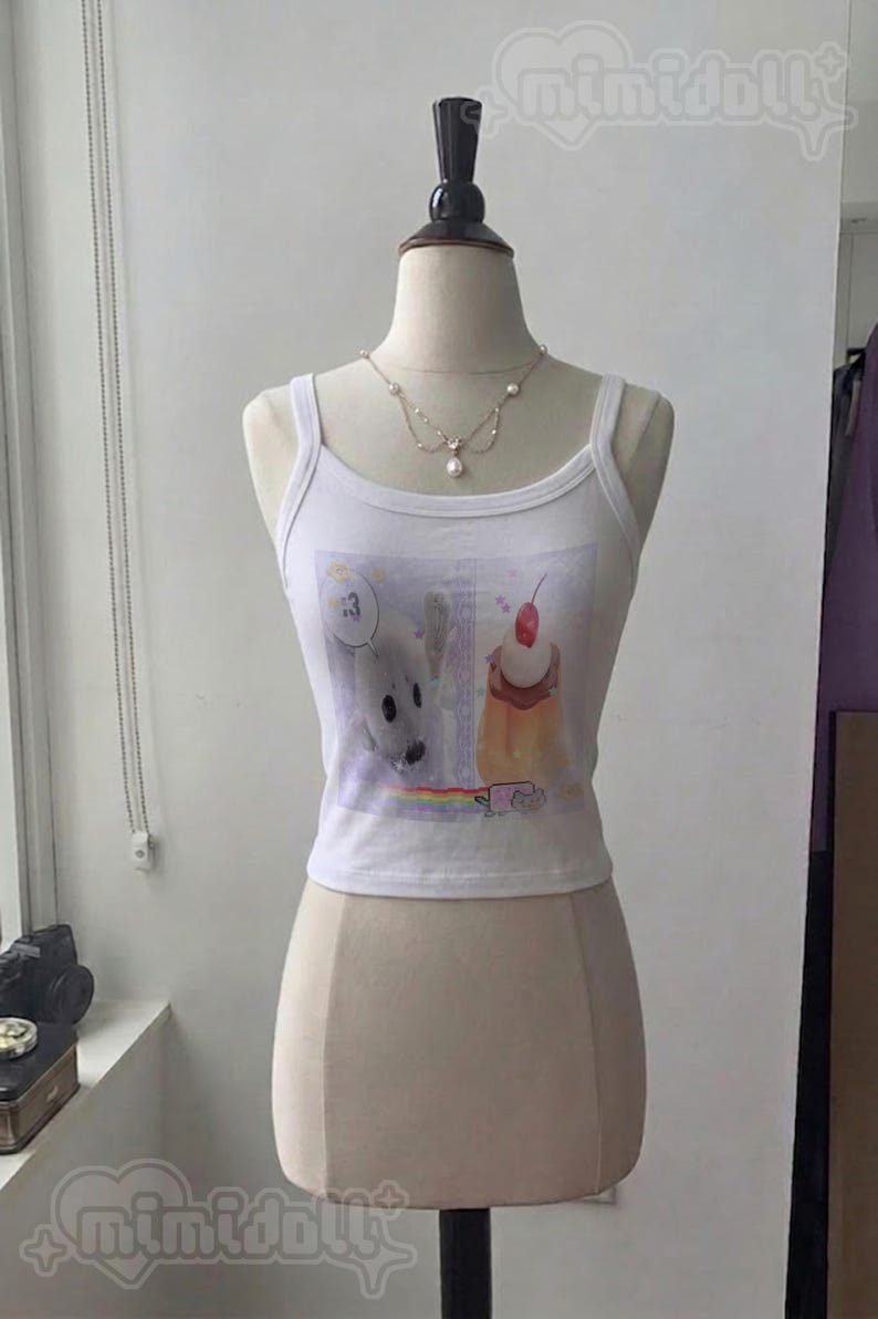 May include: A white sleeveless tank top featuring a graphic print of a white seal with a speech bubble containing the number 3, a dessert topped with a cherry, and a small rainbow. The tank top is displayed on a mannequin wearing a pearl necklace.
