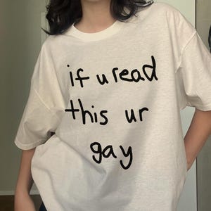 May include: White t-shirt with the handwritten text "if u read this ur gay" in black. The shirt has a crew neck and short sleeves. The fabric appears to be a soft, casual material.