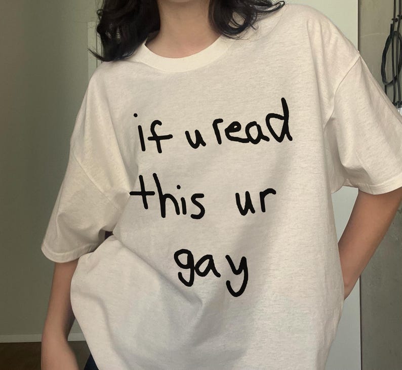 May include: White t-shirt with the handwritten text "if u read this ur gay" in black. The shirt has a crew neck and short sleeves. The fabric appears to be a soft, casual material.