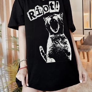 Pode incluir: T-shirt preto com um gr&aacute;fico branco de um gato a gritar e a palavra "Riot!" por cima. O texto est&aacute; numa fonte de bloco. A t-shirt tem mangas curtas e um corte folgado. A ilustra&ccedil;&atilde;o do gato &eacute; branca.