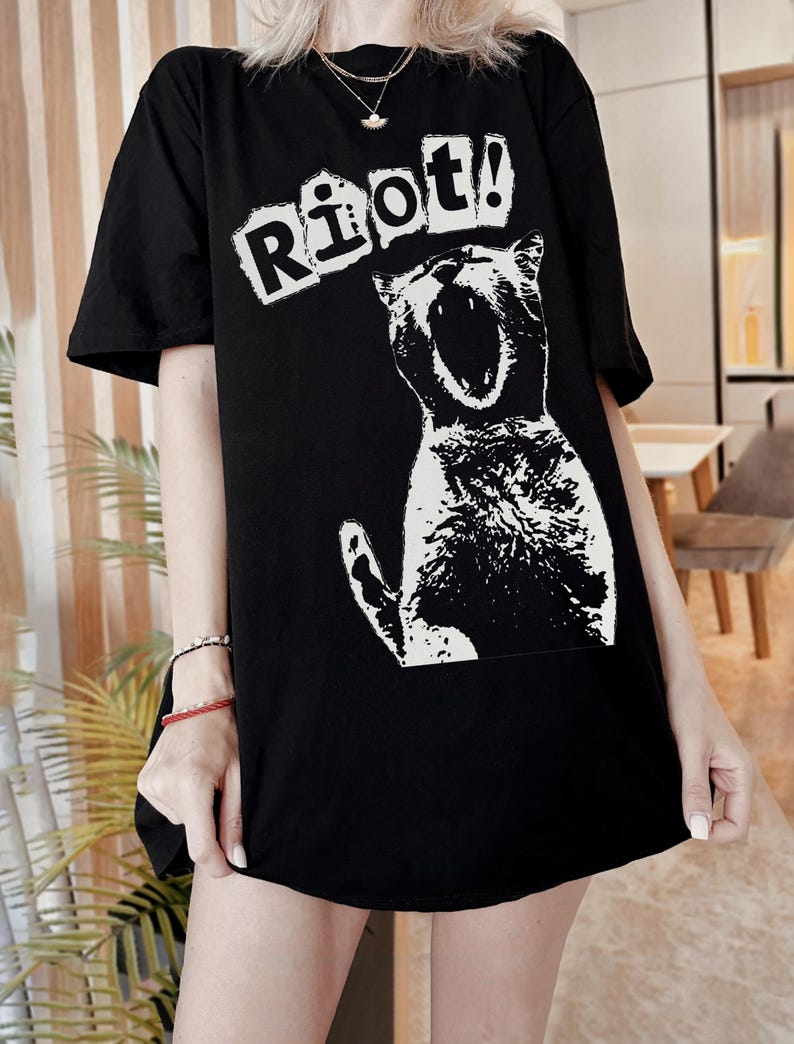 Pode incluir: T-shirt preto com um gr&aacute;fico branco de um gato a gritar e a palavra "Riot!" por cima. O texto est&aacute; numa fonte de bloco. A t-shirt tem mangas curtas e um corte folgado. A ilustra&ccedil;&atilde;o do gato &eacute; branca.