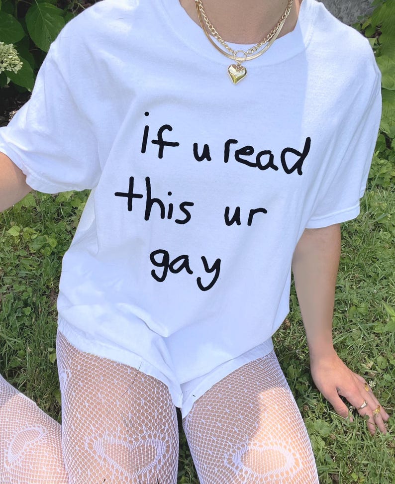 May include: White t-shirt with the handwritten text "if u read this ur gay" in black. The shirt is paired with white shorts and fishnet stockings. A gold necklace with a heart pendant is visible.
