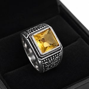 May include: A silver-toned ring featuring a large, square-cut yellow gemstone set in the center. The band is crafted with intricate, embossed floral-like patterns along the sides, providing a textured appearance to the metal surface.