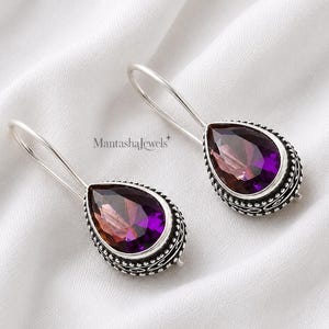 Peut inclure: Une paire de boucles d'oreilles pendantes avec des pierres en forme de poire aux facettes dégradées violettes et orange, serties dans des montures en argent oxydé ornées de perles et de longs crochets incurvés. Le texte MantashaJewels est visible.