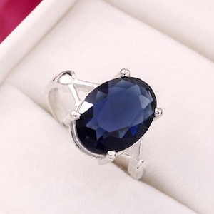 May include: A silver-toned ring featuring a large, oval-cut, deep blue gemstone held in place by four prongs. The band of the ring has a split design that connects to the setting, showcasing a polished metallic finish and a faceted stone surface.