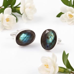 以下が含まれることがあります： 楕円形のラブラドライトストーンが付いた、シルバーのカフスボタンのペア。石は青、緑、茶色の色合いが混ざっています。カフスボタンは、白い花と緑の葉を背景に白い背景に配置されています。