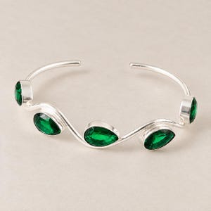 May include: A silver cuff bracelet featuring a wavy, open-ended band design. The bracelet is accented with five pear-shaped, faceted emerald green stones set in smooth silver bezels, spaced evenly along the undulating length of the metal band.