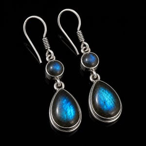 Puede incluir: Un par de pendientes colgantes de plata con dos piedras de labradorita cada uno. La piedra superior es circular y la inferior tiene forma de lágrima. Ambas piedras muestran una iridiscencia azul vibrante en monturas de plata pulida.
