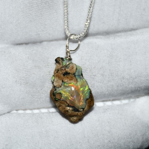 Può includere: Un ciondolo in opale naturale con una catena d'argento. L'opale ha una forma grezza e organica con un mix di tonalità marroni, beige e verdi. L'opale mostra lampi di colore iridescente, tra cui rosso, arancione e giallo.