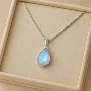 May include: A silver necklace featuring a teardrop-shaped moonstone pendant with a blue iridescent glow. The pendant is suspended from a delicate silver box chain, showcasing a smooth, polished metal setting that frames the translucent gemstone.
