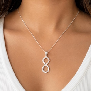 Puede incluir: Un collar de tono plateado que presenta un colgante con el símbolo de infinito vertical con una superficie texturizada y facetada. El colgante cuelga de una cadena fina y delicada de tono plateado que descansa sobre la clavícula.