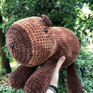 May include: A handmade brown crochet stuffed animal with a rounded body, small ears, and black button eyes. The plush toy has long, dangling legs and is held up against a backdrop of green foliage.