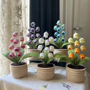 May include: A collection of handmade crochet lily of the valley plants in various colors, including pink, purple, white, blue, yellow, and orange. Each plant is potted in a beige basket. The flowers and leaves are detailed, creating a charming, decorative display.
