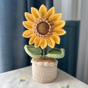 Op de afbeelding: Een gehaakte zonnebloem in een beige pot. De zonnebloem heeft heldergele bloemblaadjes en een bruin hart. Groene bladeren en stelen ondersteunen de bloem. De pot heeft een geschulpte rand en staat op een lichtgekleurd oppervlak.