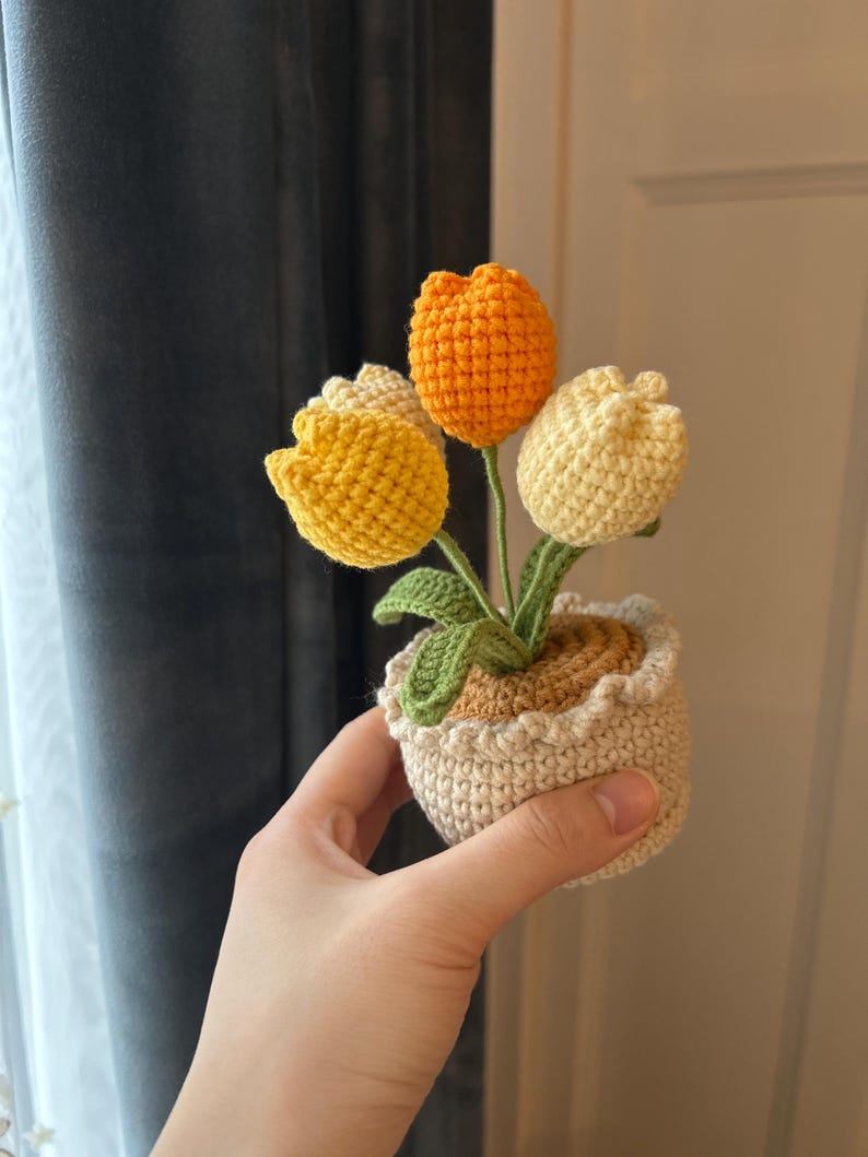 May include: A crocheted tulip arrangement in a small pot. The flowers are yellow, orange, and cream-colored, with green leaves. The pot is beige with a ruffled edge. The arrangement is held in a hand.