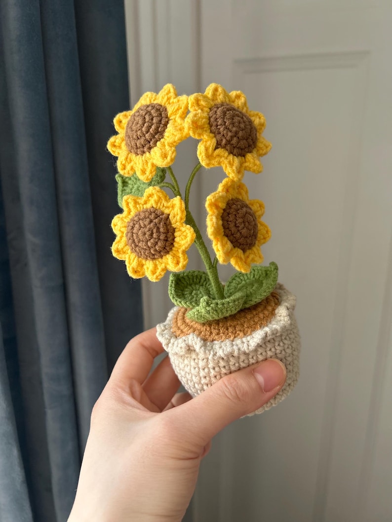 May include: A crocheted sunflower arrangement in a small pot. The sunflowers have yellow petals and brown centers. The pot is beige with a ruffled edge. The leaves are green. The arrangement is held in a hand.