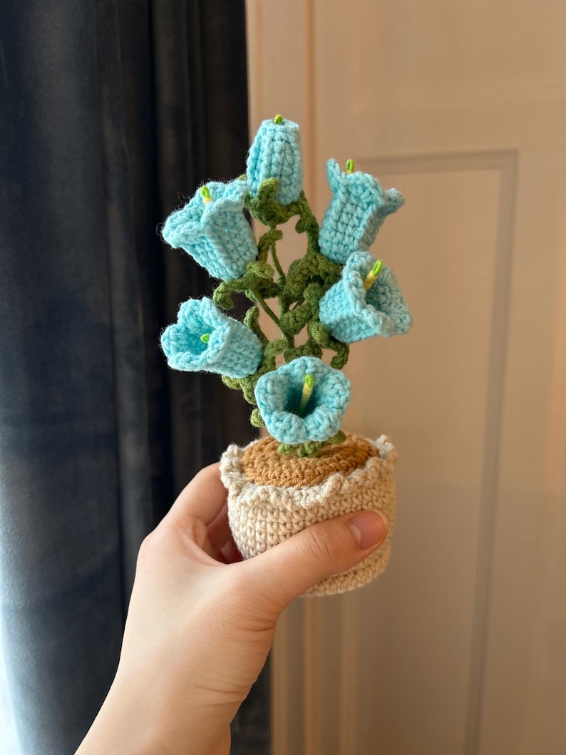 May include: A handmade crochet flower arrangement featuring light blue bell-shaped flowers with green stems and leaves. The flowers are in a beige pot with a ruffled edge. The arrangement is held in a hand.