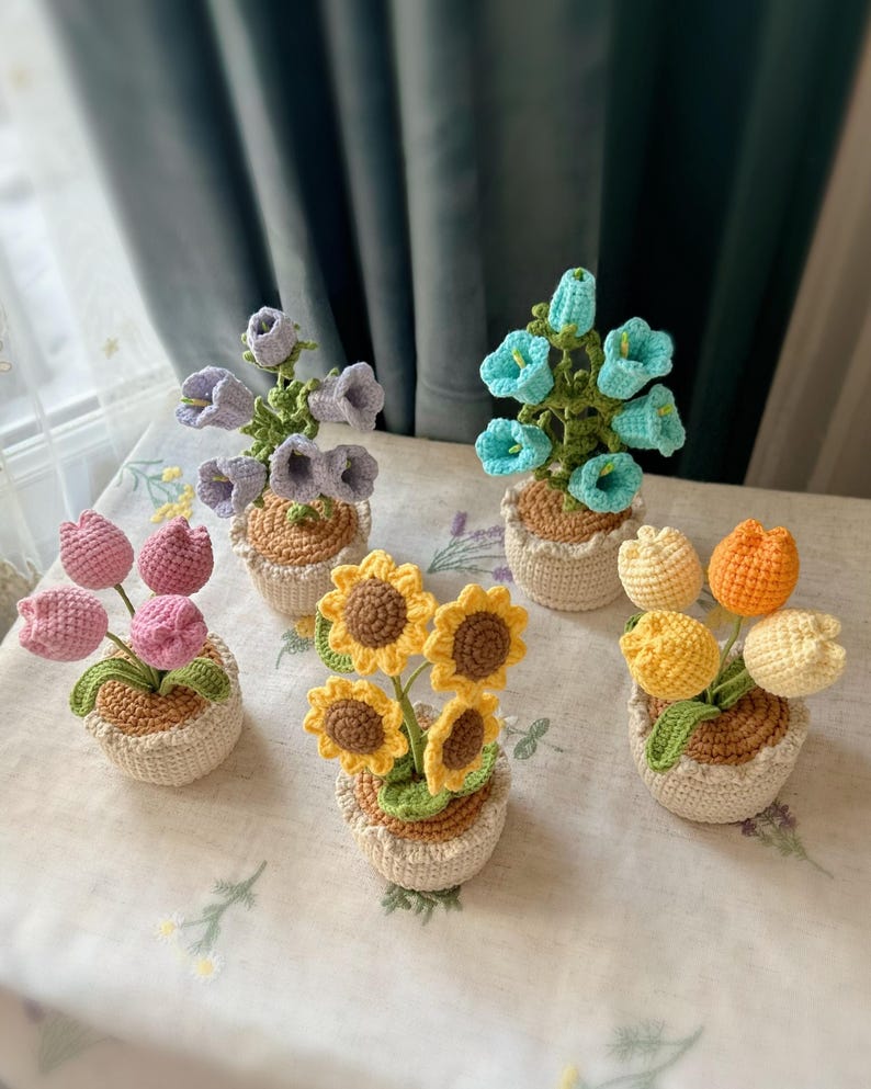 May include: A collection of handmade crochet flower arrangements. The arrangements include tulips, sunflowers, and bell-shaped flowers in various colors, such as pink, yellow, and blue. Each flower is potted in a small, woven basket.