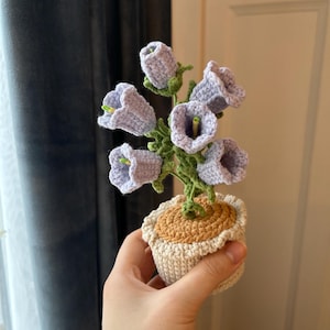 May include: A handmade crochet flower arrangement featuring lavender bell-shaped flowers with green stems and leaves. The flowers are in a small, off-white pot with a tan top. The arrangement is held in a hand.