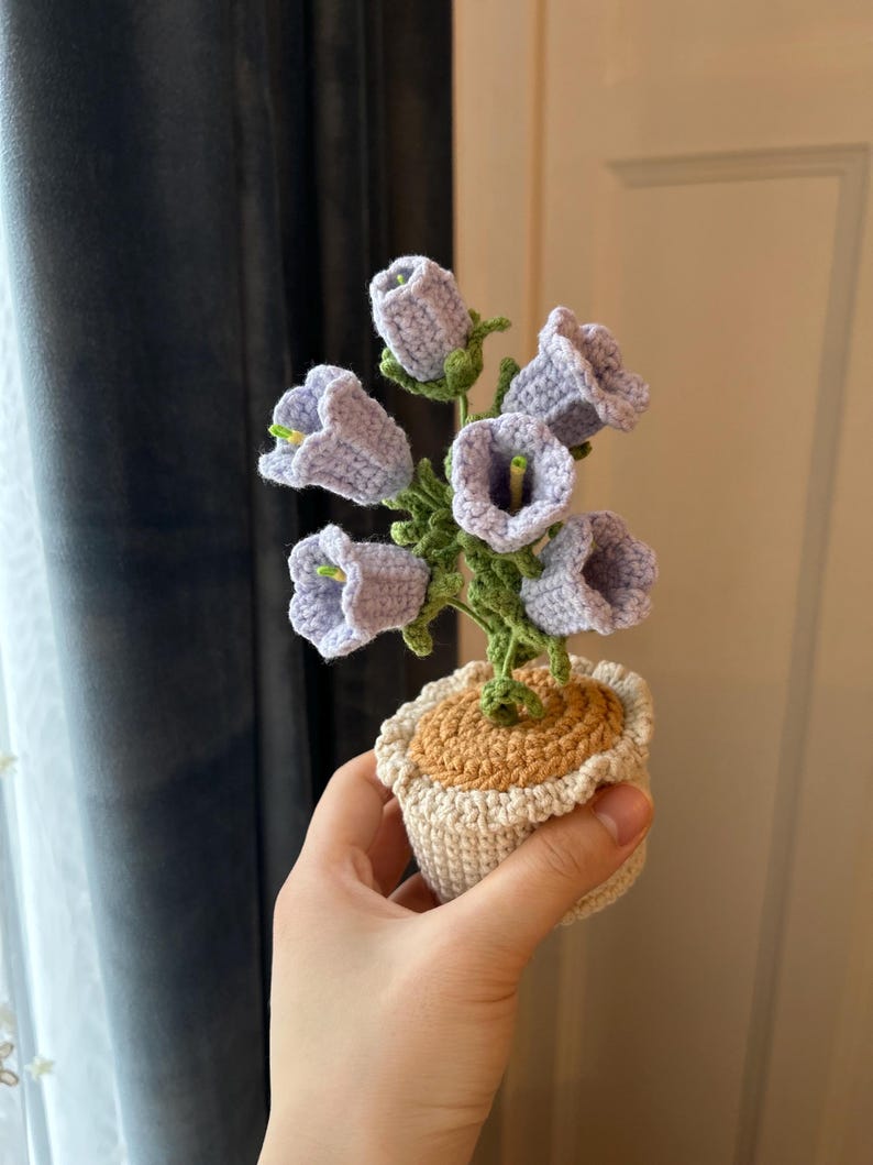 May include: A handmade crochet flower arrangement featuring lavender bell-shaped flowers with green stems and leaves. The flowers are in a small, off-white pot with a tan top. The arrangement is held in a hand.