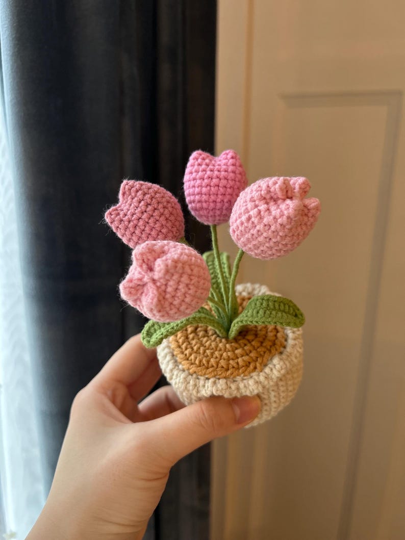 May include: A handmade crochet tulip arrangement in a small pot. The tulips are various shades of pink, with green stems and leaves. The pot is beige and tan. The craftwork is held in a hand.