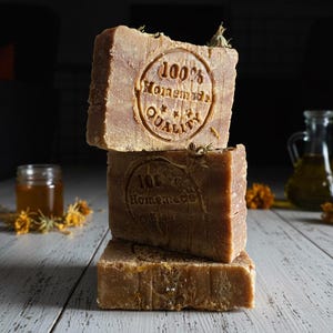 Può includere: Tre saponette quadrate marroni impilate, con le parole "100% Homemade Quality" stampate sopra. Il sapone è su una superficie di legno bianco, con un piccolo barattolo e una bottiglia di vetro sullo sfondo.