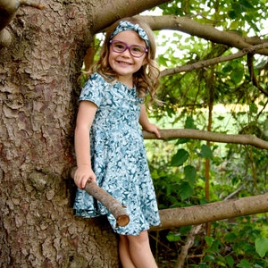 May include: A short-sleeved dress featuring a teal and white floral pattern, paired with a matching knotted fabric headband. The dress has a round neckline and a flared skirt. The ensemble is completed by purple-framed eyeglasses with clear lenses.