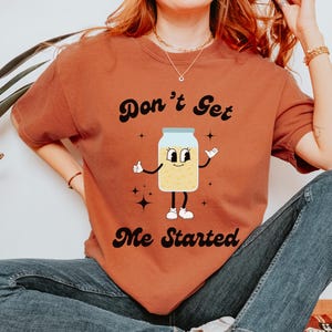 May include: A rust-colored t-shirt with a cartoon illustration of a jar with a face and the text "Don't Get Me Started".