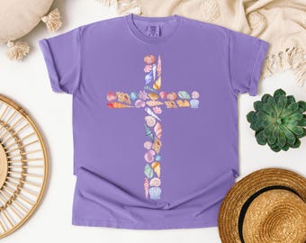 Könnte beinhalten: Ein lilafarbenes T-Shirt mit einem Kreuzdesign aus verschiedenen Muscheln in Rosa, Orange, Blau und Weiß. Das kurzärmelige Shirt besteht aus einem weichen Material und eignet sich für den Freizeitgebrauch.