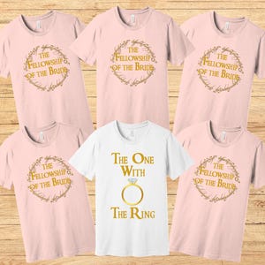 May include: Set of six pink t-shirts with gold text and graphics. The shirts feature the text "The Fellowship of the Bride" and "The One with the Ring".