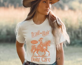 May include: White t-shirt with a graphic of a woman riding a horse. The text "Trail Hair Don't Care" is printed on the shirt.