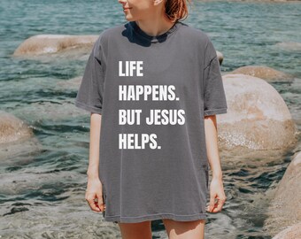 May include: A gray t-shirt with the text "LIFE HAPPENS. BUT JESUS HELPS." in white block letters. The shirt is worn by a person standing near water and rocks.