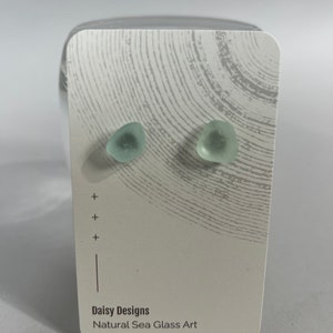 Może przedstawiać: Para kolczyków ze szkła morskiego na białej kartce z tekstem "Daisy Designs Natural Sea Glass Art".