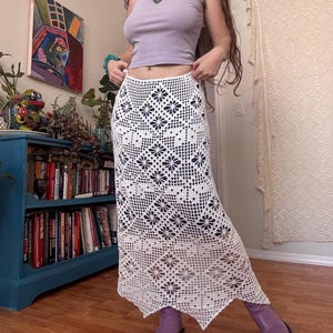 May include: A white crochet skirt with a floral pattern. The skirt is long and has a scalloped hemline. The skirt is paired with a light purple tank top and purple boots. The skirt is handmade and has a bohemian style.