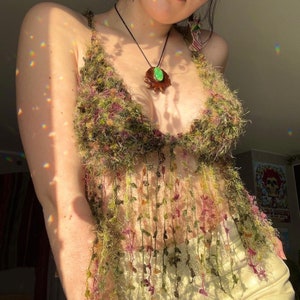 Puede incluir: Un top de crochet verde y marrón con flecos. El top se lleva sobre un top blanco sin mangas. La persona que lleva el top también lleva un collar con un colgante de piedra verde.